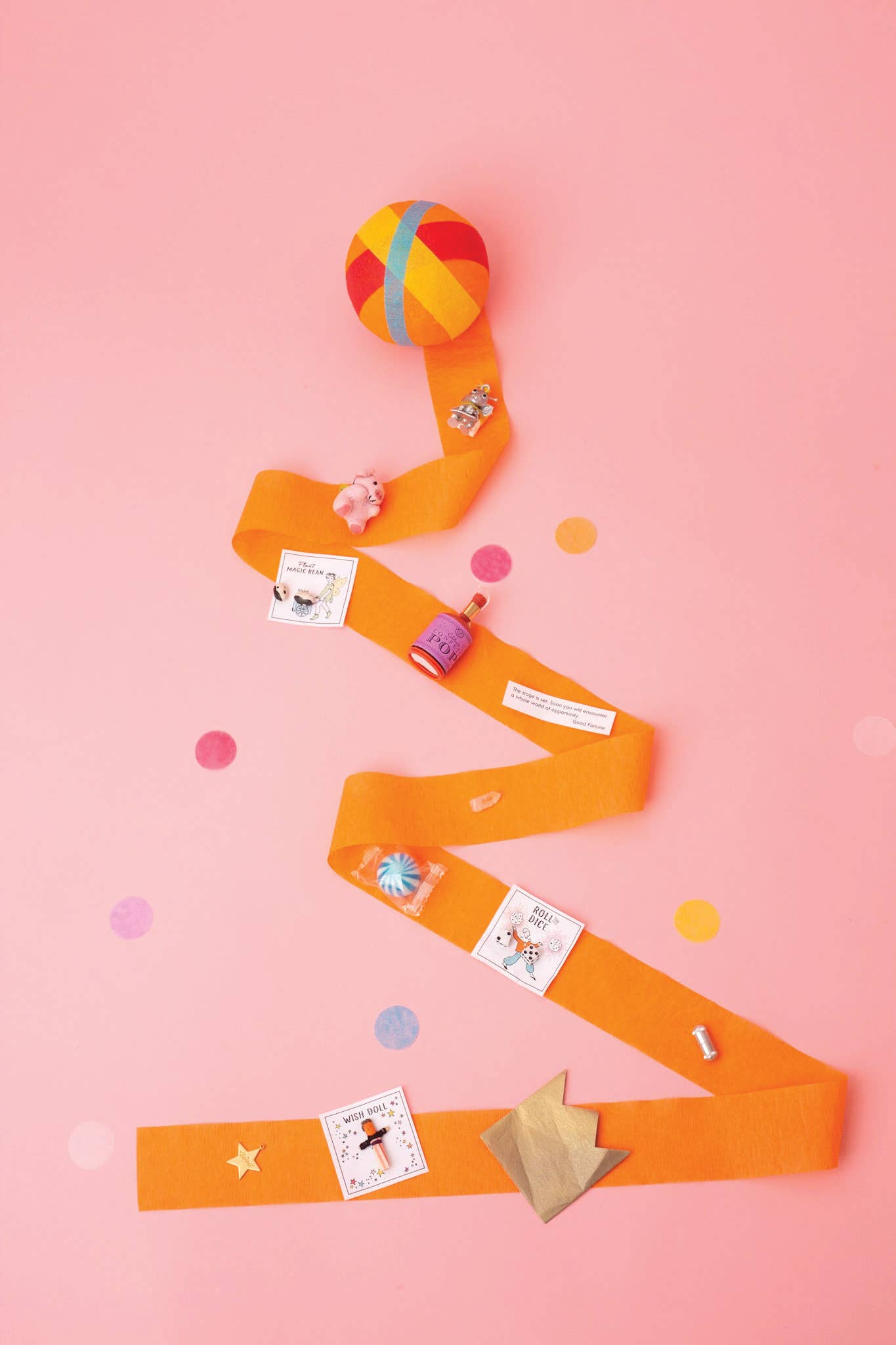 A surprise ball unraveled with its contents, mini confetti bottle, crown, toys and a joke, on a pink background.