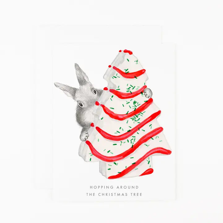 A white greeting card featuring a watercolor illustration of a gray rabbit peeking around a white Christmas tree decorated with red icing-like stripes and green sprinkles. The text at the bottom reads &