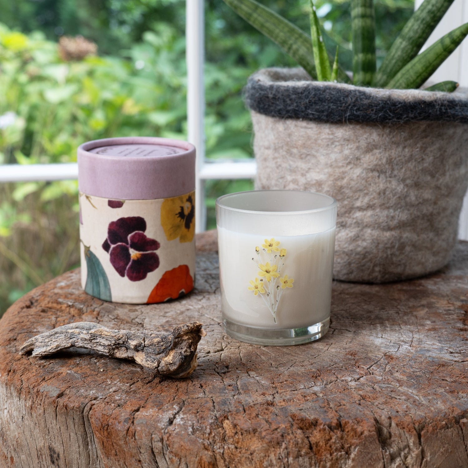Candle with flower design on a wooden surface with a plant in the background.