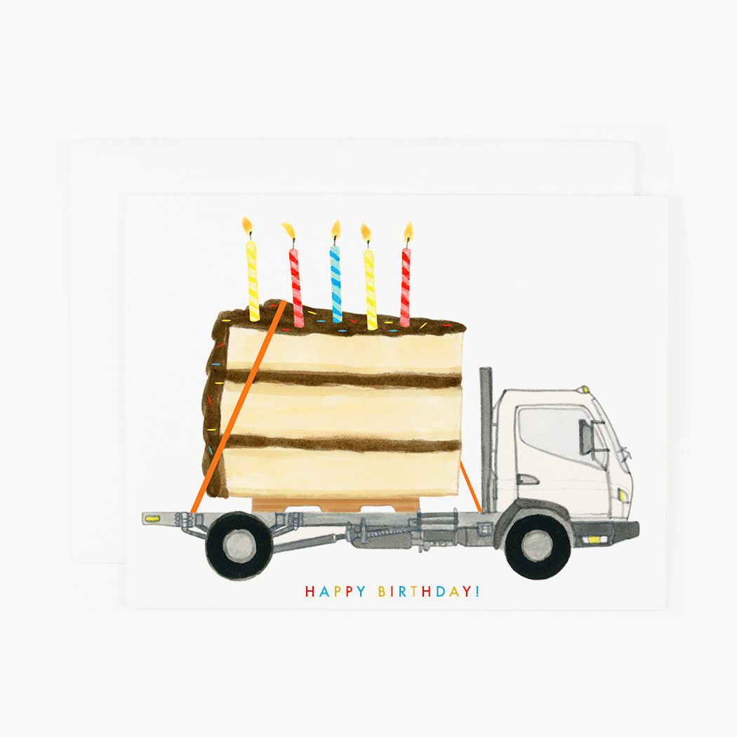 A greeting card with a gigantic piece of cake with sprinkles and candles on a flatbed truck with colorful text that reads &quot;Happy birthday&quot;.