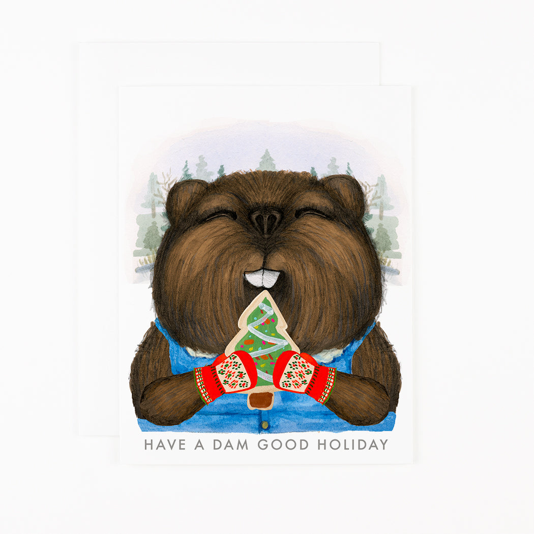 A Dear Hancock card featuring a hand-drawn beaver with a vest and mittens, eating a tree-shaped cookie and text that reads &quot;Have a dam good holiday&quot; underneath.