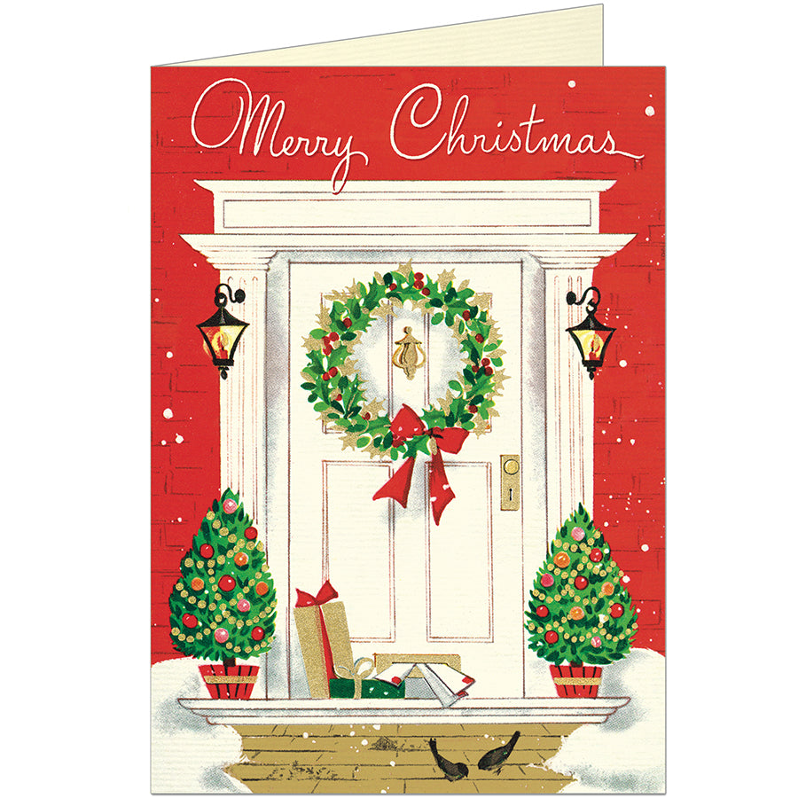 A Christmas greeting card featuring an illustrated white front door with a green wreath and red bow, set against a red brick wall. The card reads &