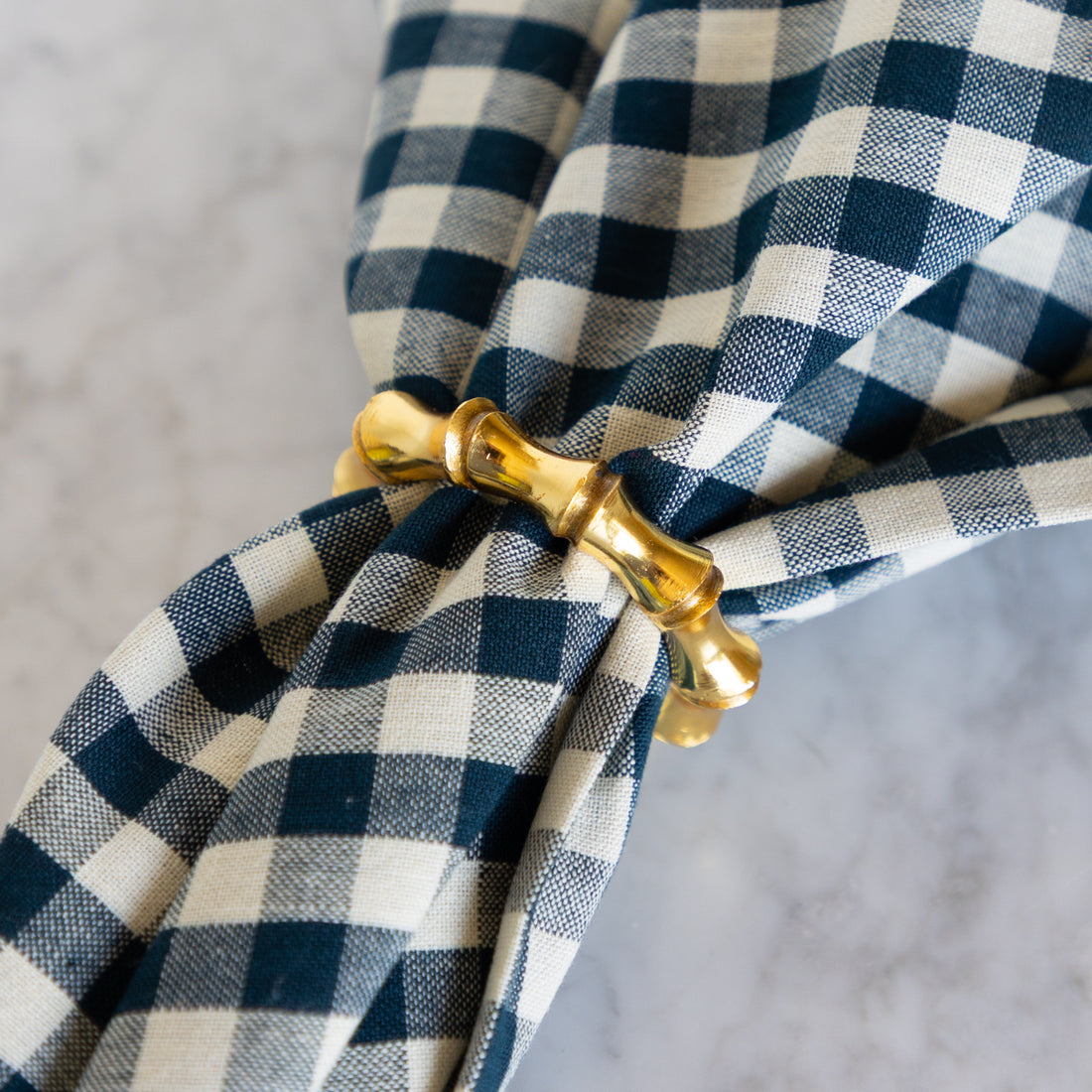 Blue and white checkered napkin with a gold napkin ring on a gray background.
