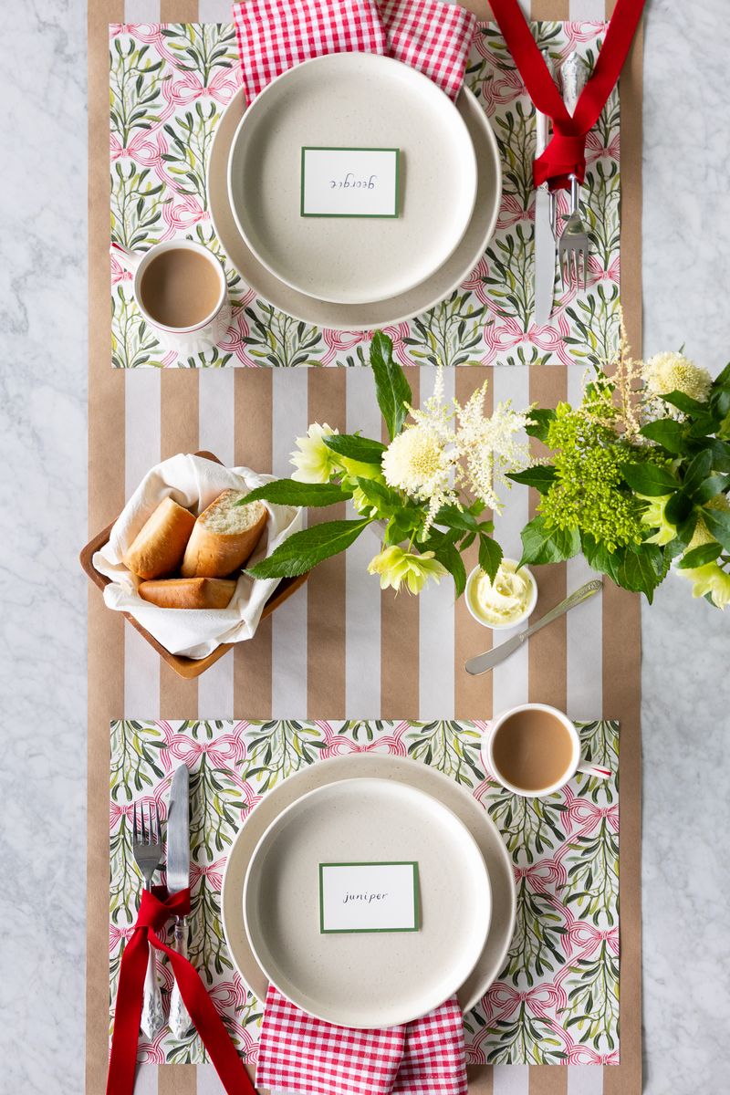 A holiday tablescape featuring mistletoe placemats on a kraft and white striped runner.
