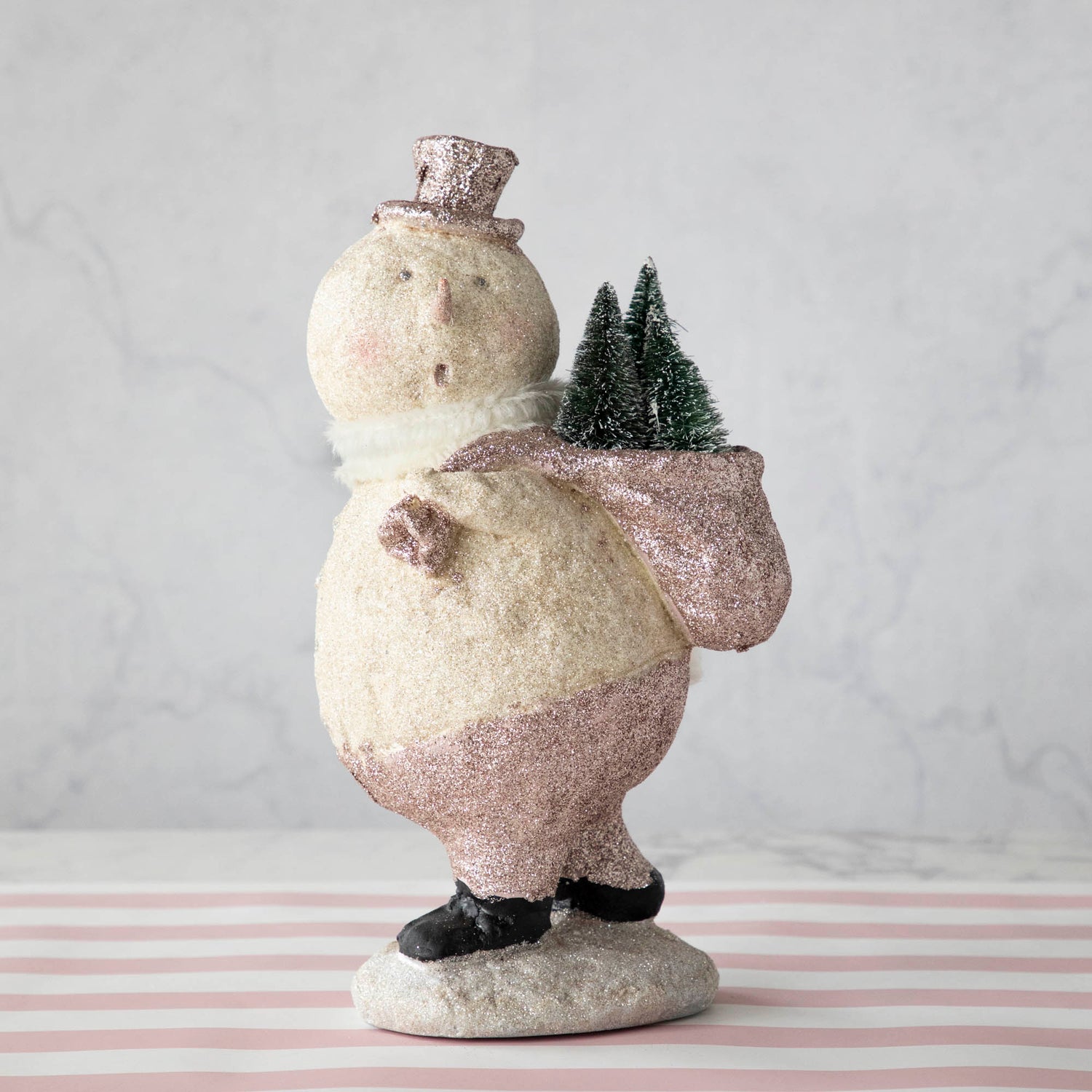 A Sparkle Snowman figurine carrying a sack of trees on its back while standing on a pile of snow.