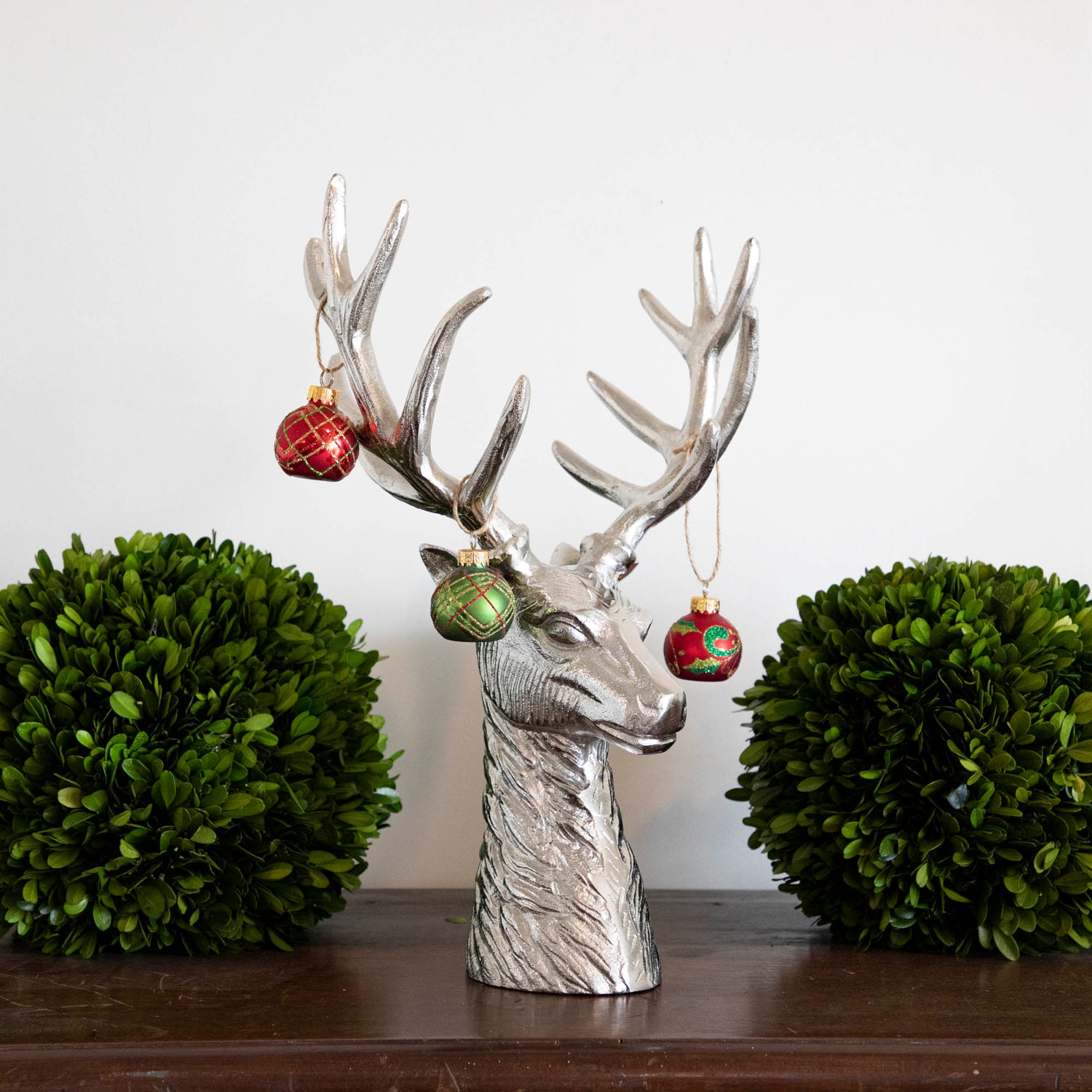 An aluminum Reindeer Bust with ornaments hanging on its antlers, between two boxwood balls on mantle.