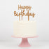 A white frosted cake with a gold glitter acrylic Meri Meri Happy Birthday Cake Toppers Set of 2 on a pink stand.