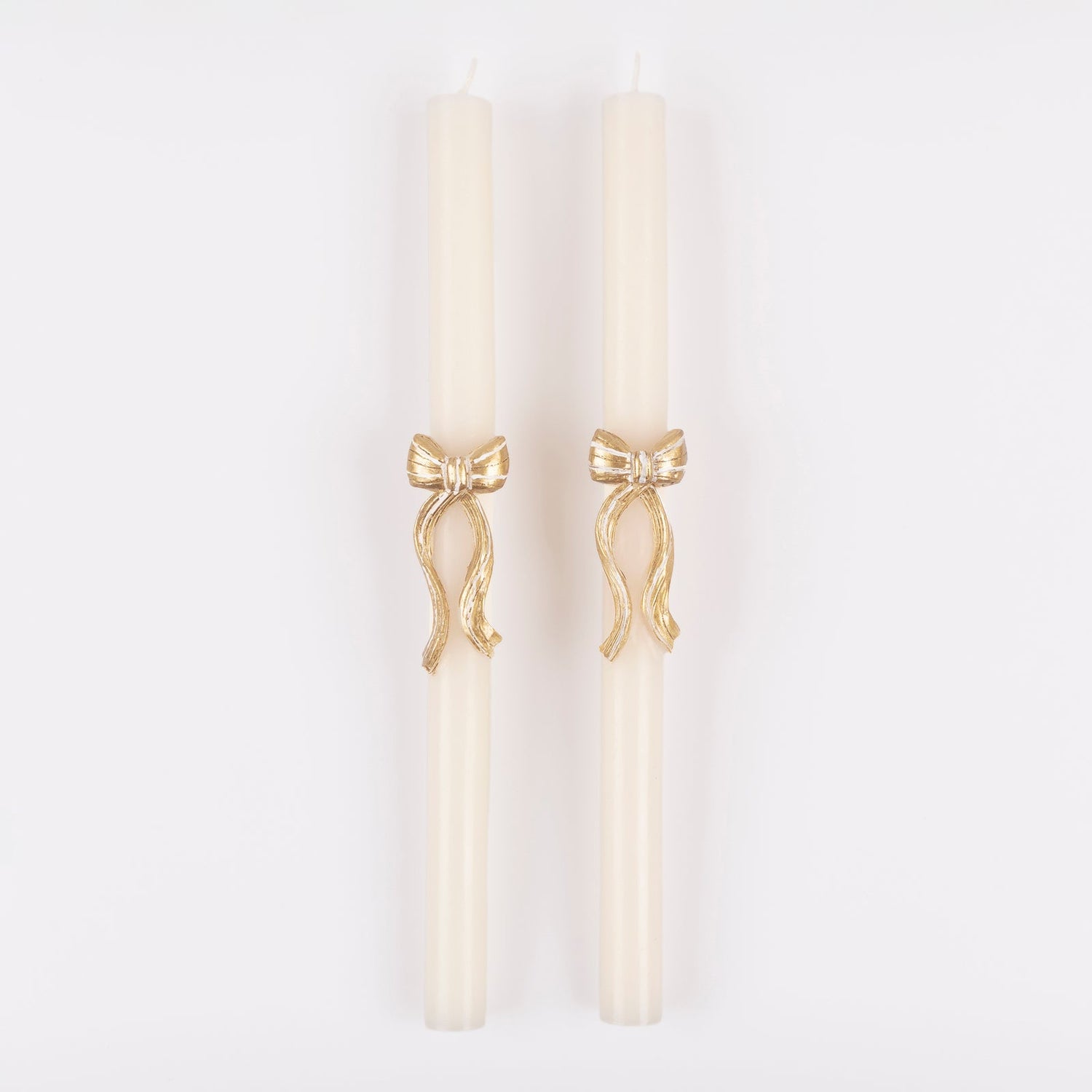 Two white taper candles with gold bow embellishments on a white background. 
