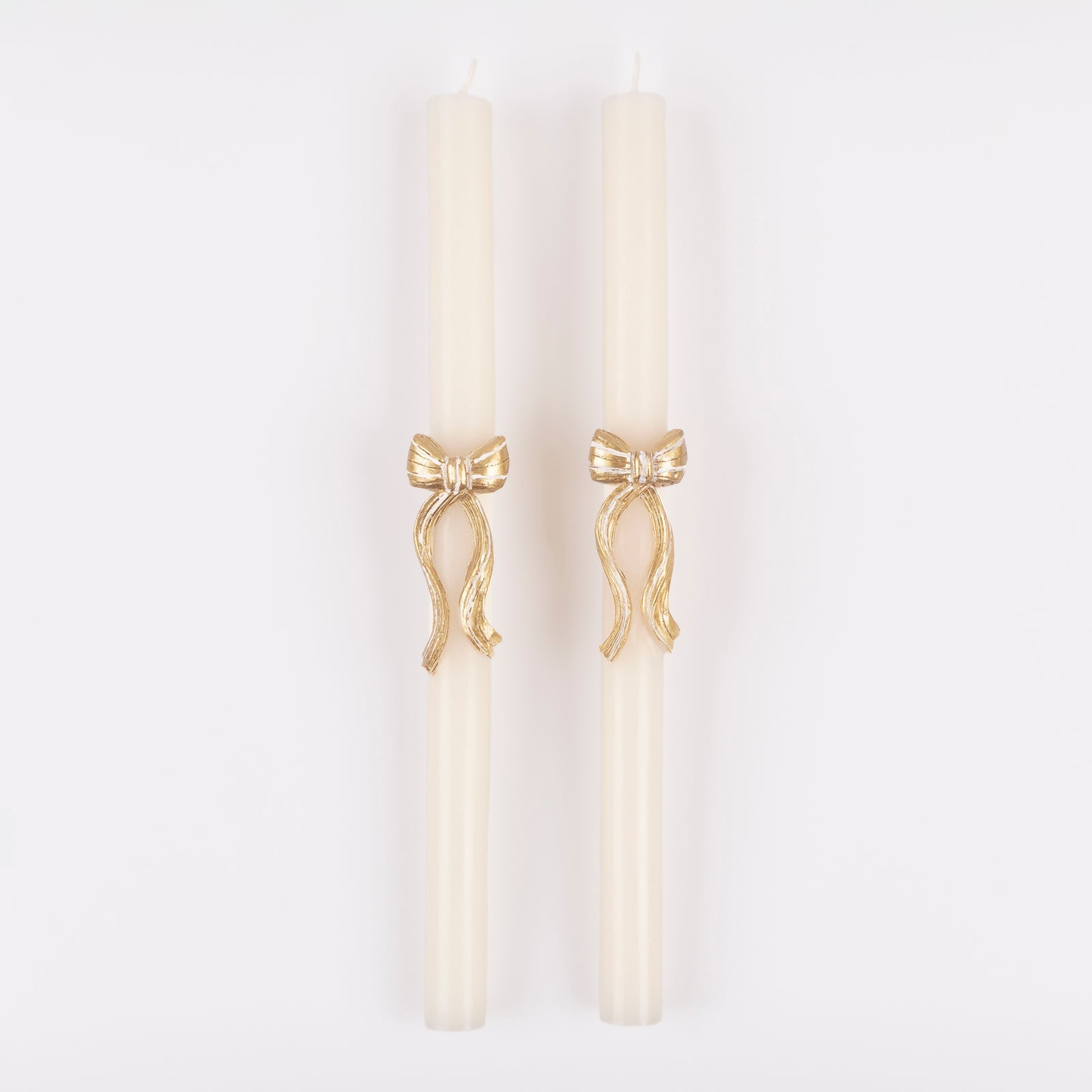 Two white taper candles with gold bow embellishments on a white background. 