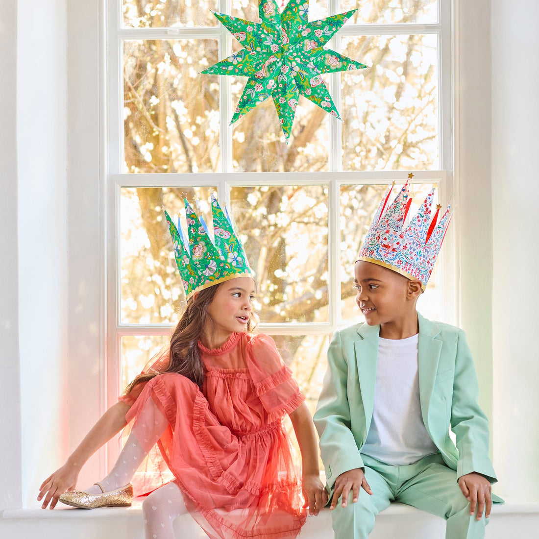 Two children wearing festive crowns sitting in front of a window with decorative lights.