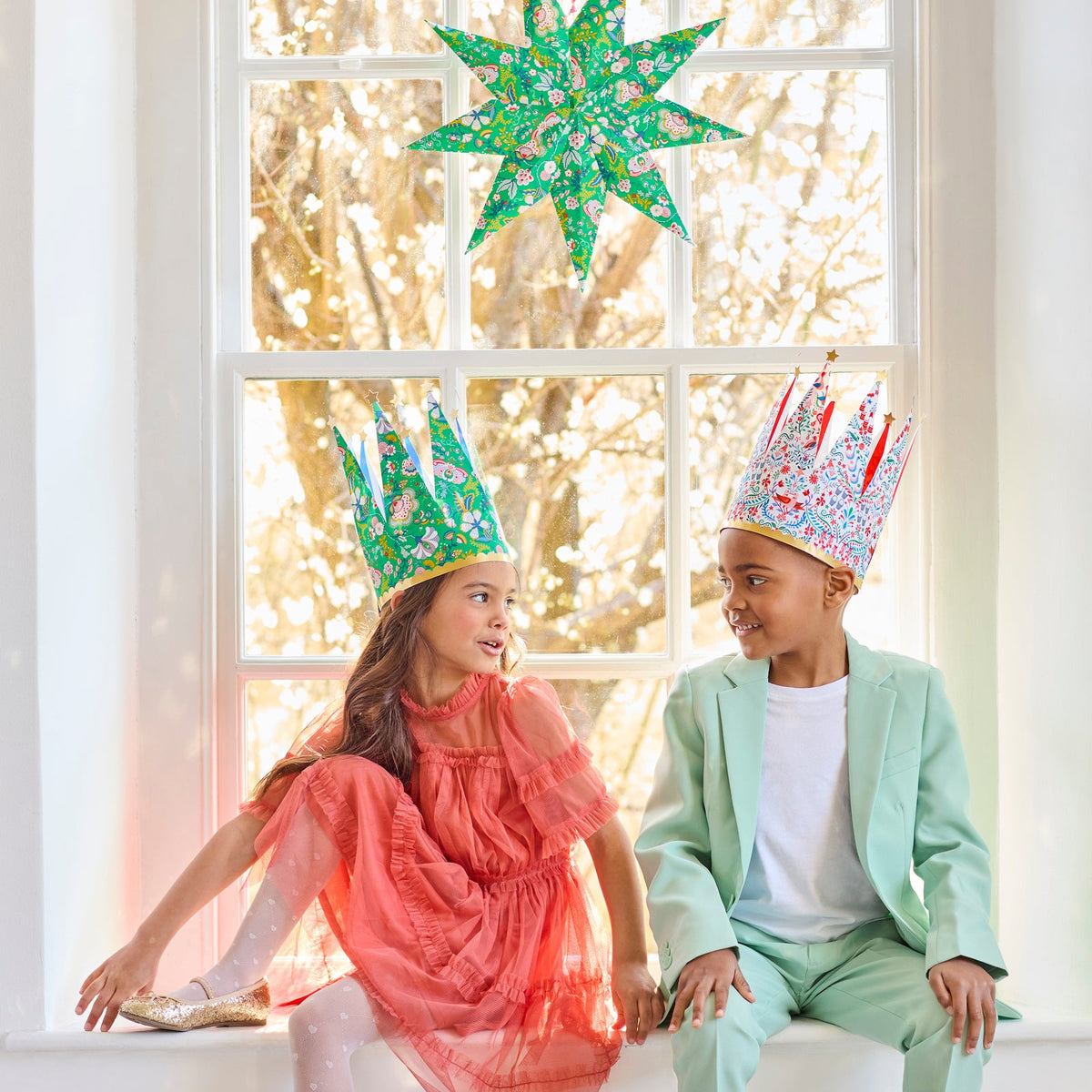 Two children wearing festive crowns sitting in front of a window with decorative lights.