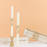 Two white candles with gold bow embellishments in gold holders on a peach background.
