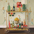A square cocktail napkin featuring a painting of a fully stocked vintage bar cart in front of a mermaid-patterned wallpaper. 