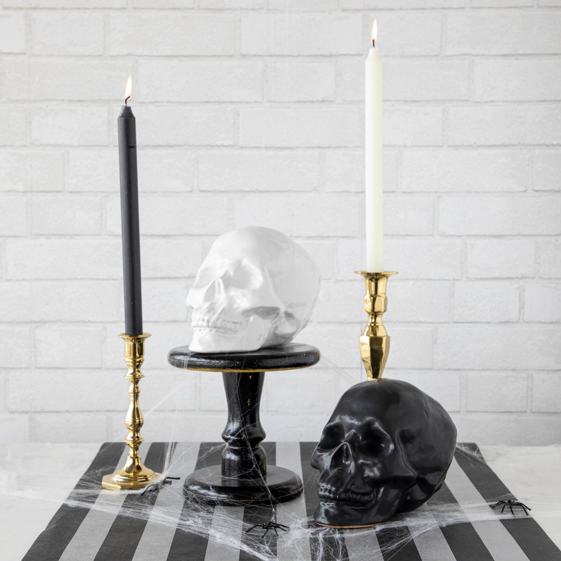 Two HomArt ceramic Skull Heads on a striped tablecloth flanked by lit candles in brass holders against a white brick background, serving as an ominous decor piece.
