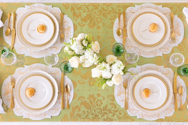 Elegant table setting with white plates, gold cutlery, and floral arrangements on a green and gold patterned table runner.