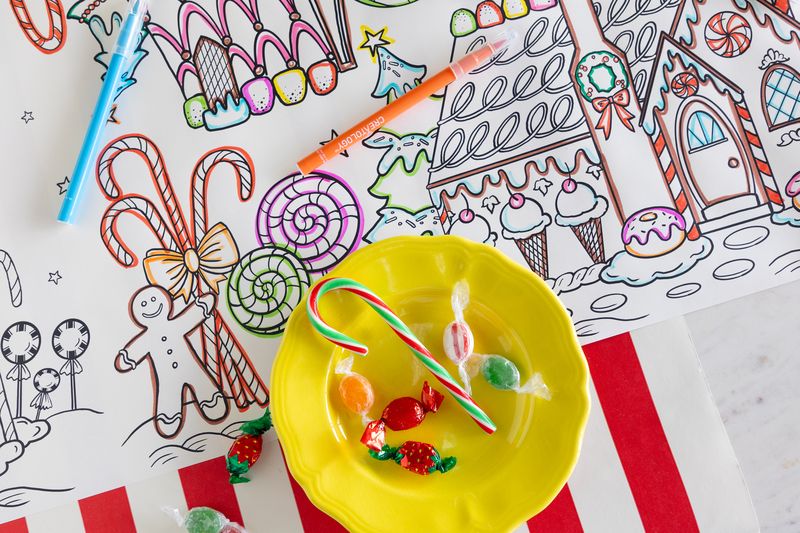 Yellow bowl with candy canes and gumdrops on a table with a paper coloring runner featuring gingerbread houses and candy-themed illustrations that are partially colored in. 