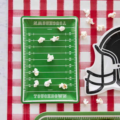 A football field with Football Field Paper Plates and a football helmet on a red and white checkered tablecloth, accompanied by game day goodies.