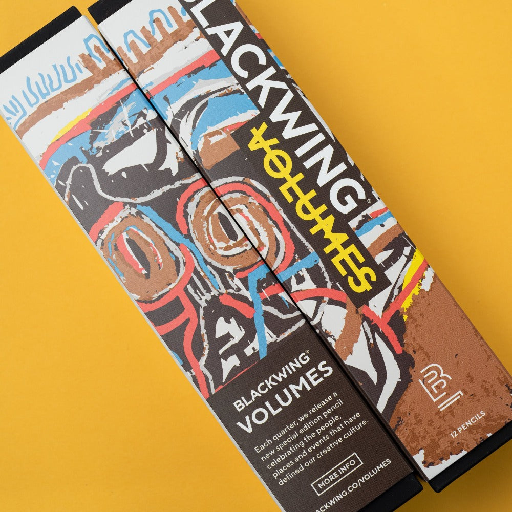 Three special-edition Blackwing Volume 57, Tribute to Jean-Michel Basquiat pencils with one sharpened and a metal sharpener on a blue background.