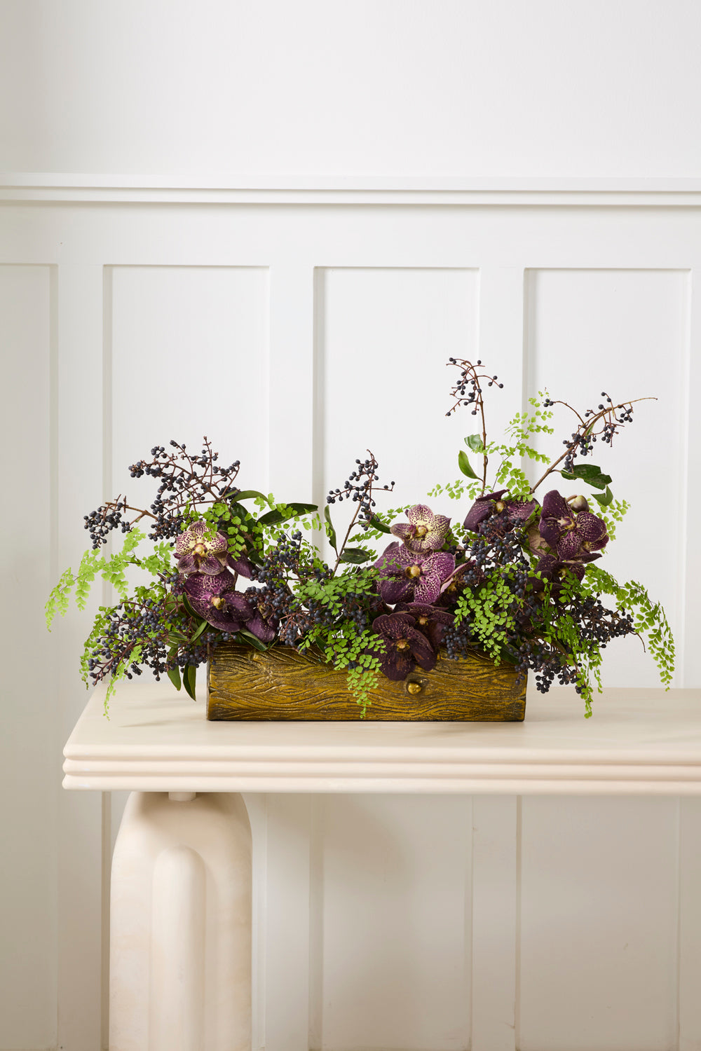 Decorative floral arrangement in a gold log planter on a white surface with a white paneled background.