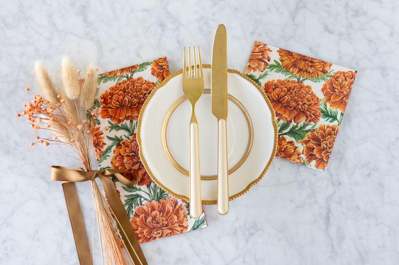 A small white and gold plate with matching cutlery sitting on Guest sized and Cocktail sized paper marigold napkins on a white marble table. 