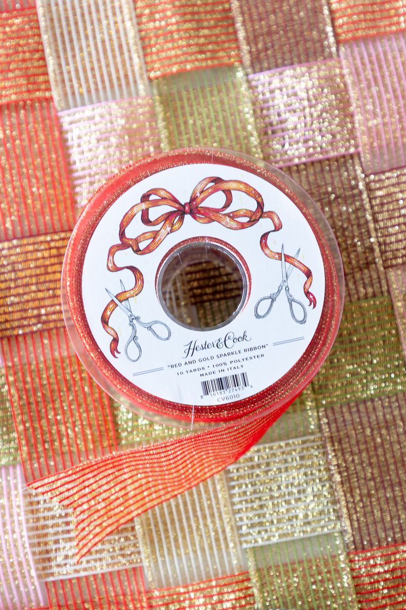 A spool of sparkly red and gold ribbon on a surface of woven sparkle ribbon in five different colors. 