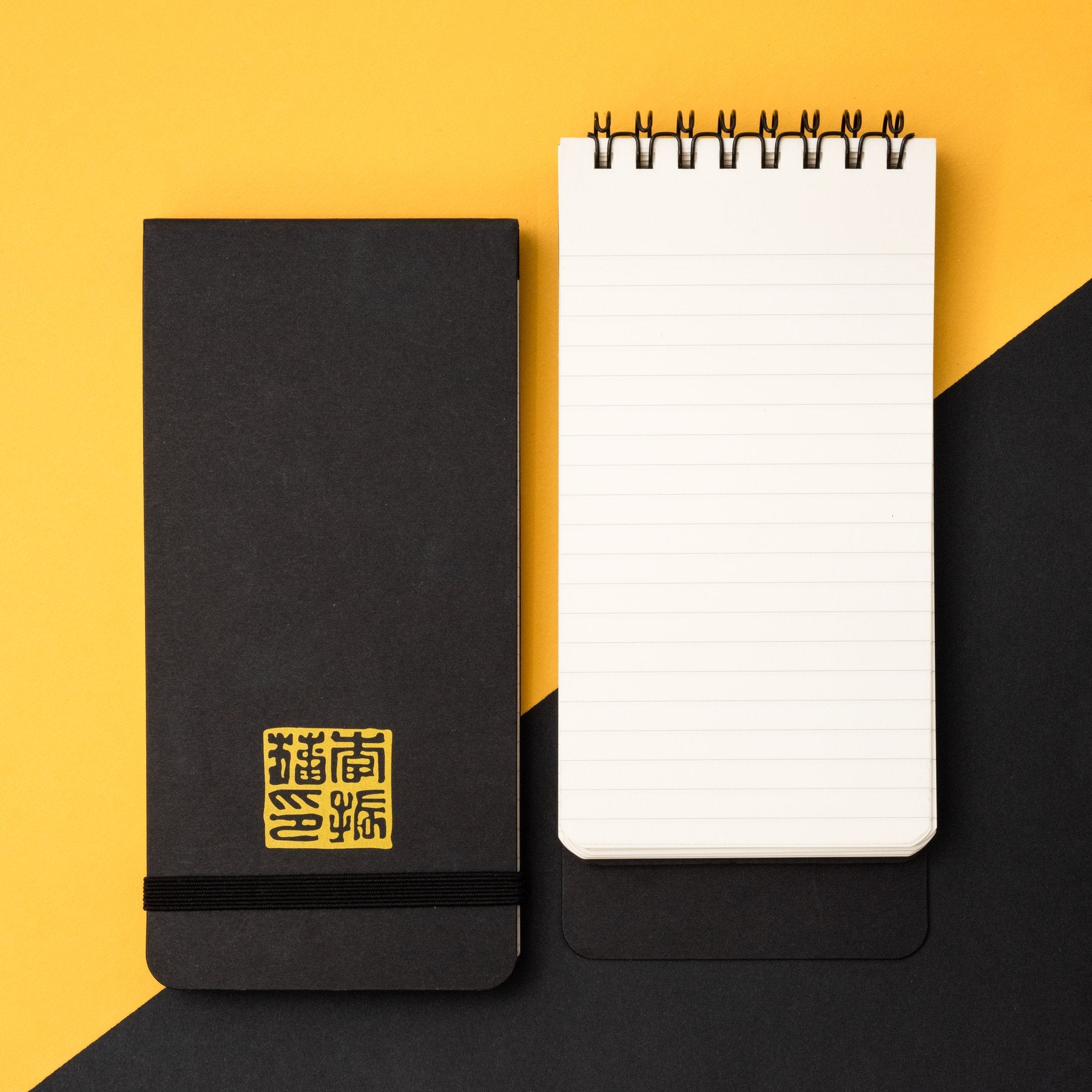 A closed Blackwing Volume 651- Tribute to Bruce Lee notebook with gold lettering next to an open spiral Reporter Pads on a dual black and yellow background.
