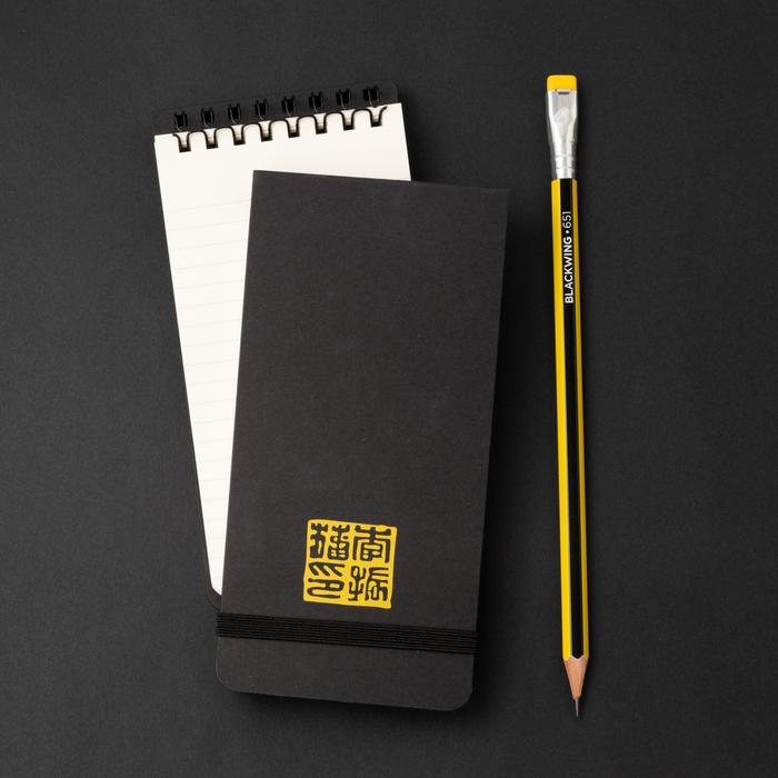 A closed Blackwing Volume 651- Tribute to Bruce Lee notebook with gold lettering next to an open spiral Reporter Pads on a dual black and yellow background.