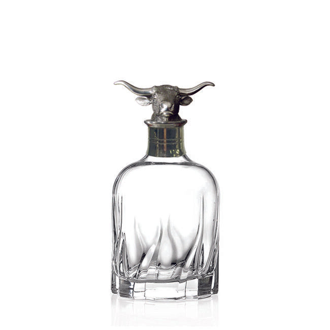 A Menagerie crystal Longhorn whisky decanter with a metallic bull head stopper accompanied by two glasses on a wooden surface, against a marble background.
