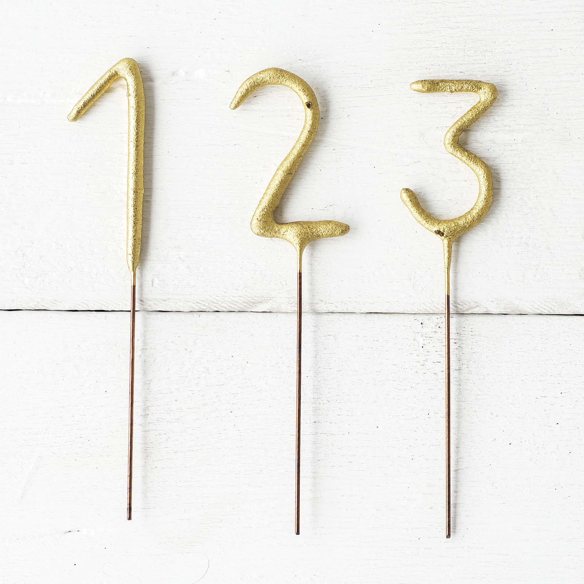 Three Tops Malibu Gold Number Cake Topper Sparklers sticks on a wooden surface, instantly transforming any celebration into an elegant affair. These 4&quot; tall decorations capture attention with a sparkling golden finish.