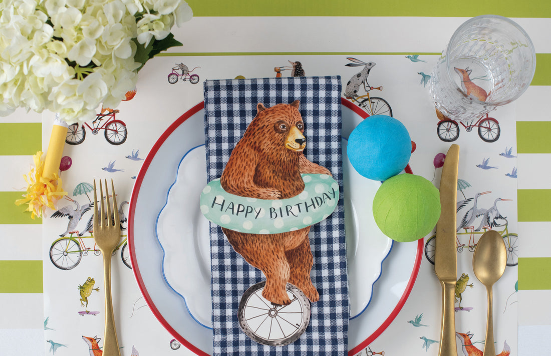 A bear illustrated balancing on a unicycle with a polka-dotted inner tube around its waist, featured as a Nice Wheels Bear Table Accent by Hester &amp; Cook.