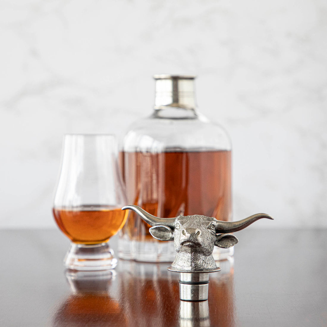A Menagerie crystal Longhorn whisky decanter with a metallic bull head stopper accompanied by two glasses on a wooden surface, against a marble background.