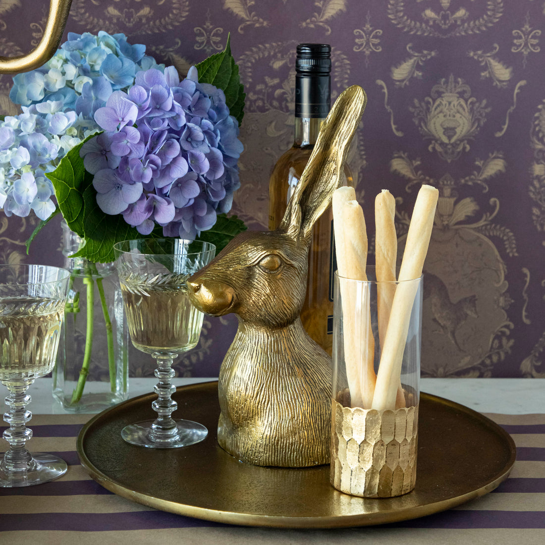 A Hare Platter by Accent Decor, with a gold bunny on a bronze platter next to a vase of flowers on a table, adding a touch of whimsy to the setting.