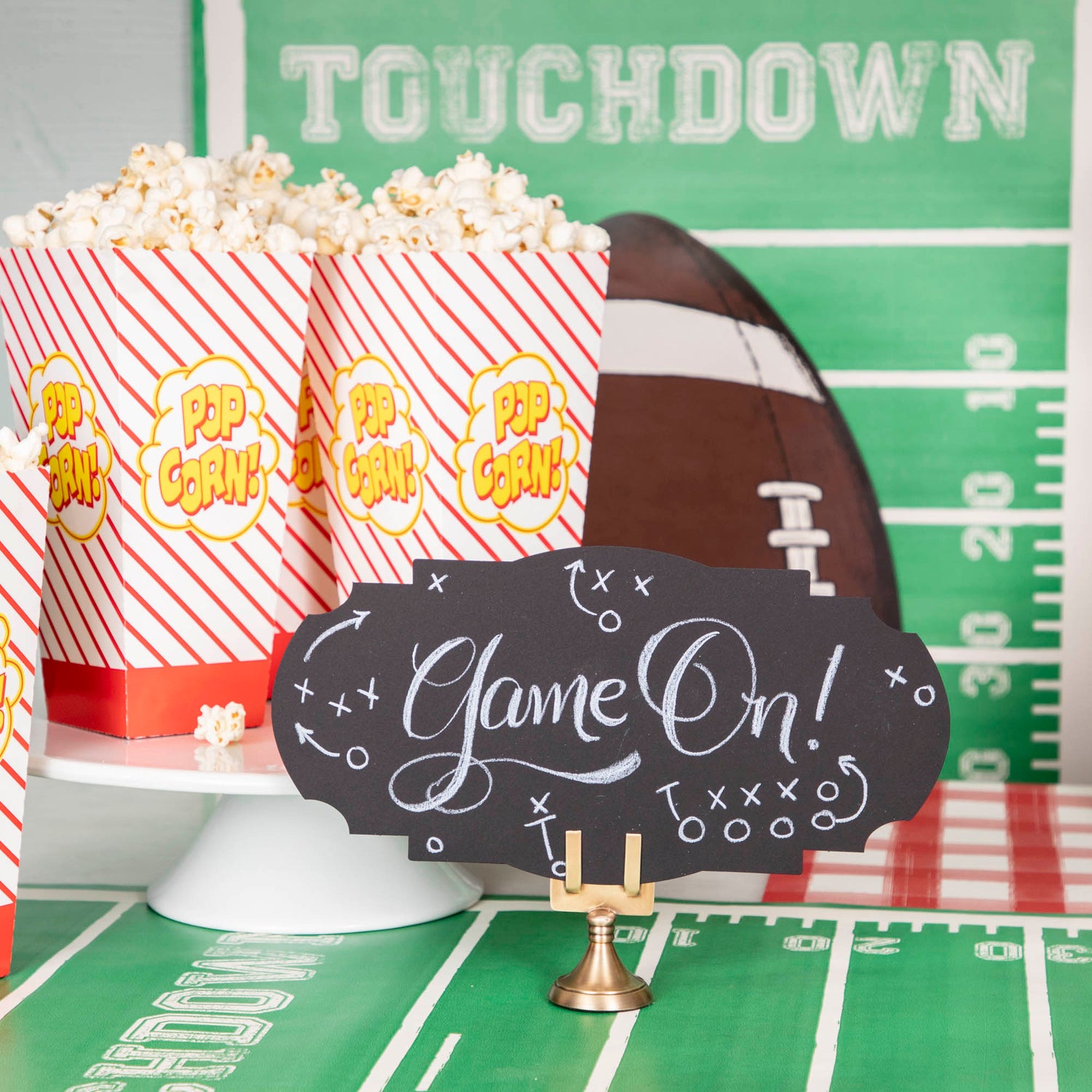 A football-themed snack table featuring a Black Frame Table Accent standing in a place card holder, with &quot;Game On!&quot; hand-written in white script along with accents of Xs and Os with arrows in the style of football strategy maps.