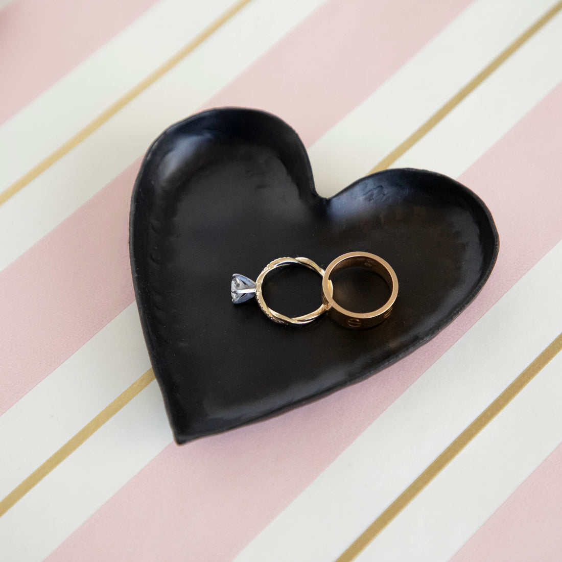 A HomArt Forged Iron Heart Tray with gold and silver rings on a copper-colored, pink, and white striped surface.