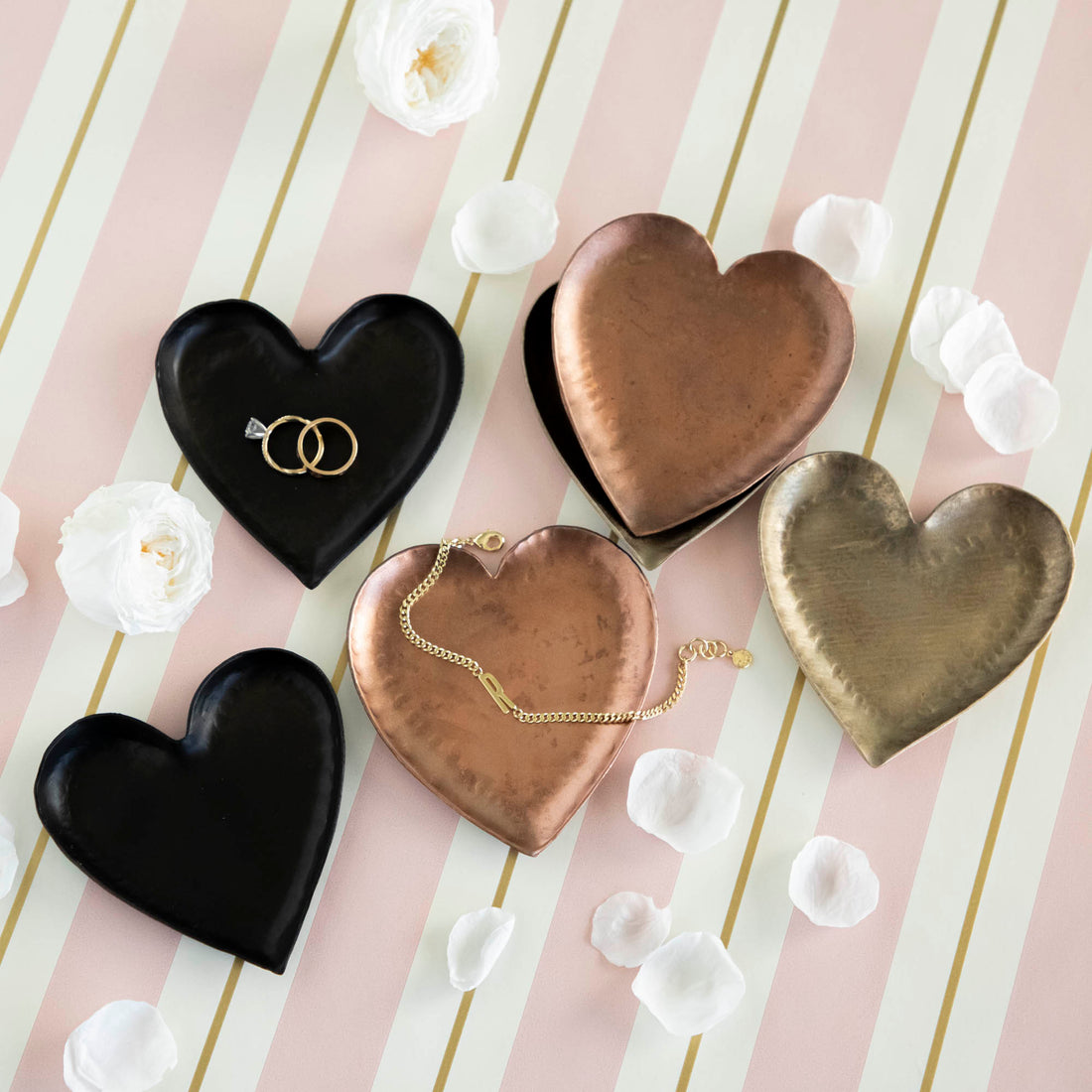 A HomArt Forged Iron Heart Tray with gold and silver rings on a copper-colored, pink, and white striped surface.