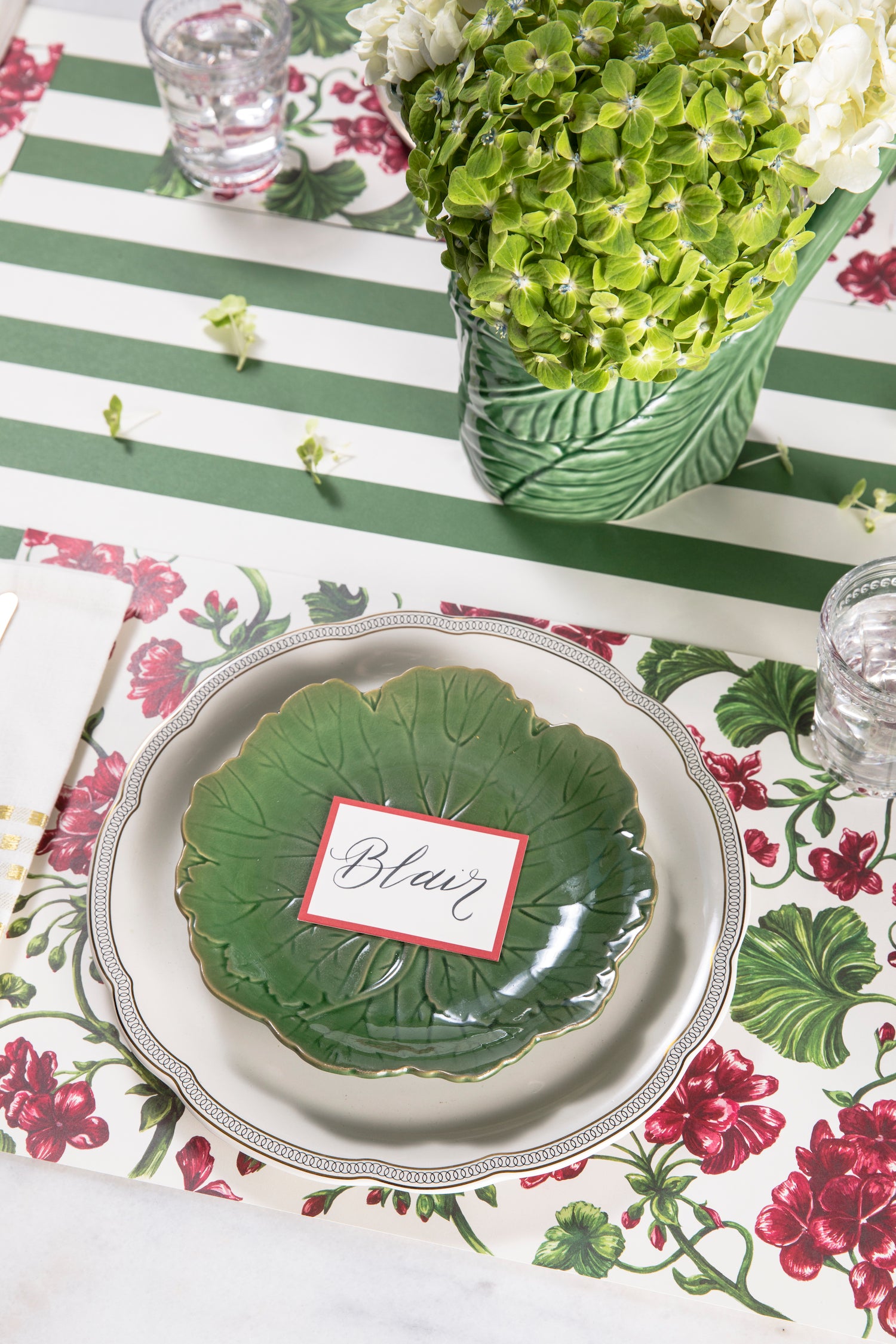 A table setting with green and white plates, Geranium Garden Placemats by Hester &amp; Cook, and flowers.