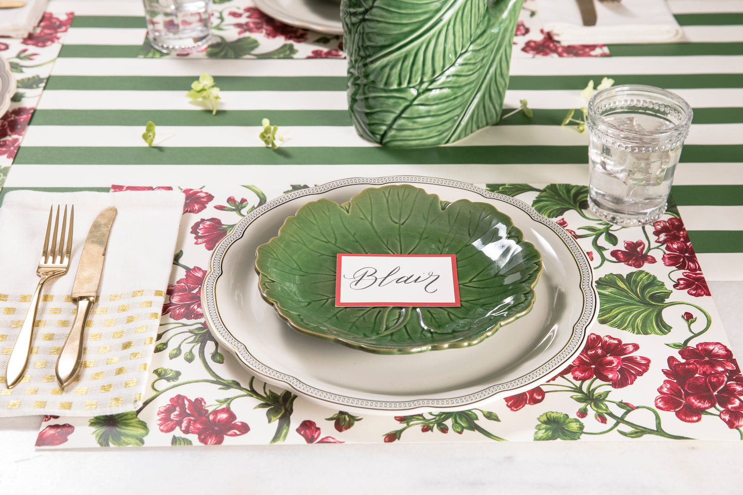 A garden-themed table setting with green and white plates and napkins, adorned with Geranium Garden Placemats from Hester &amp; Cook featuring leaves and vines.