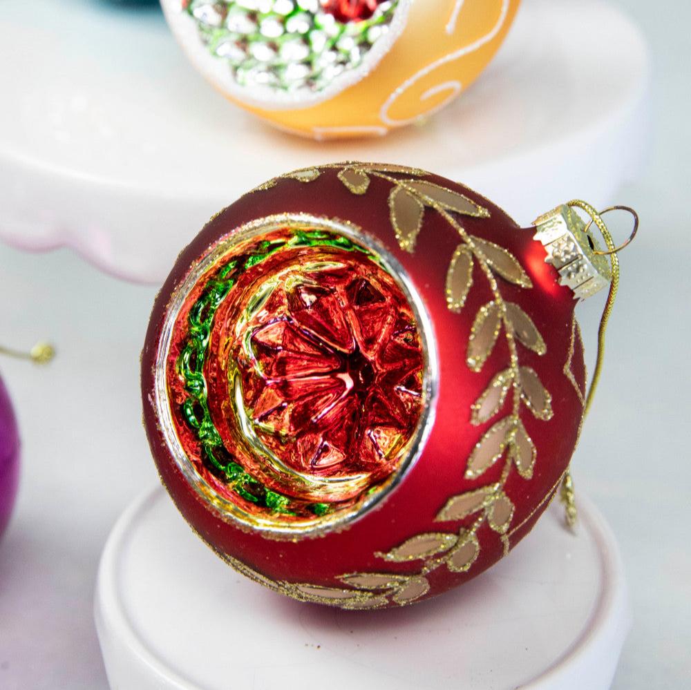 A close up of a red Reflector ornament showing its intricate design of a garland of leaves adorned with glitter wrapping around the ornament, and colorful inside.