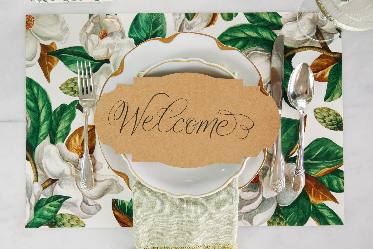 An elegant floral place setting featuring a Kraft Frame Table Accent resting on the plate with &quot;Welcome&quot; written in beautiful script.