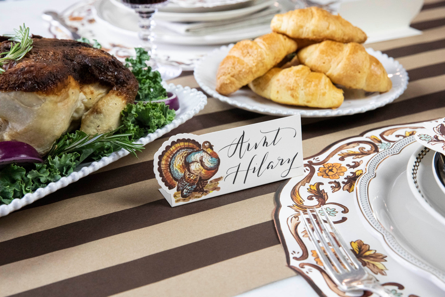 Close-up of a Thanksgiving Turkey Place Card labeled &quot;Aunt Hilary&quot; standing next to the plate of an elegant place setting.