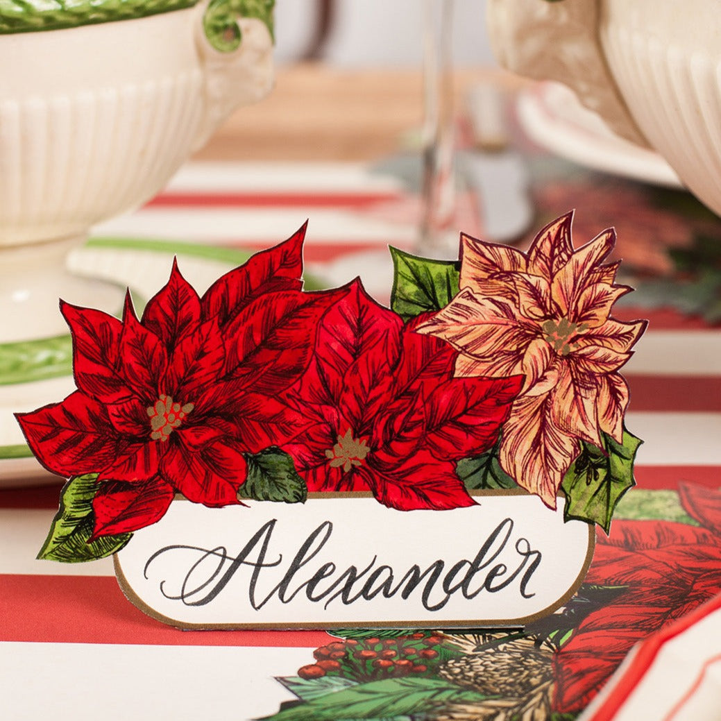 A poinsettia Place Card labeled &quot;Alexander&quot; standing on a festive holiday table setting.