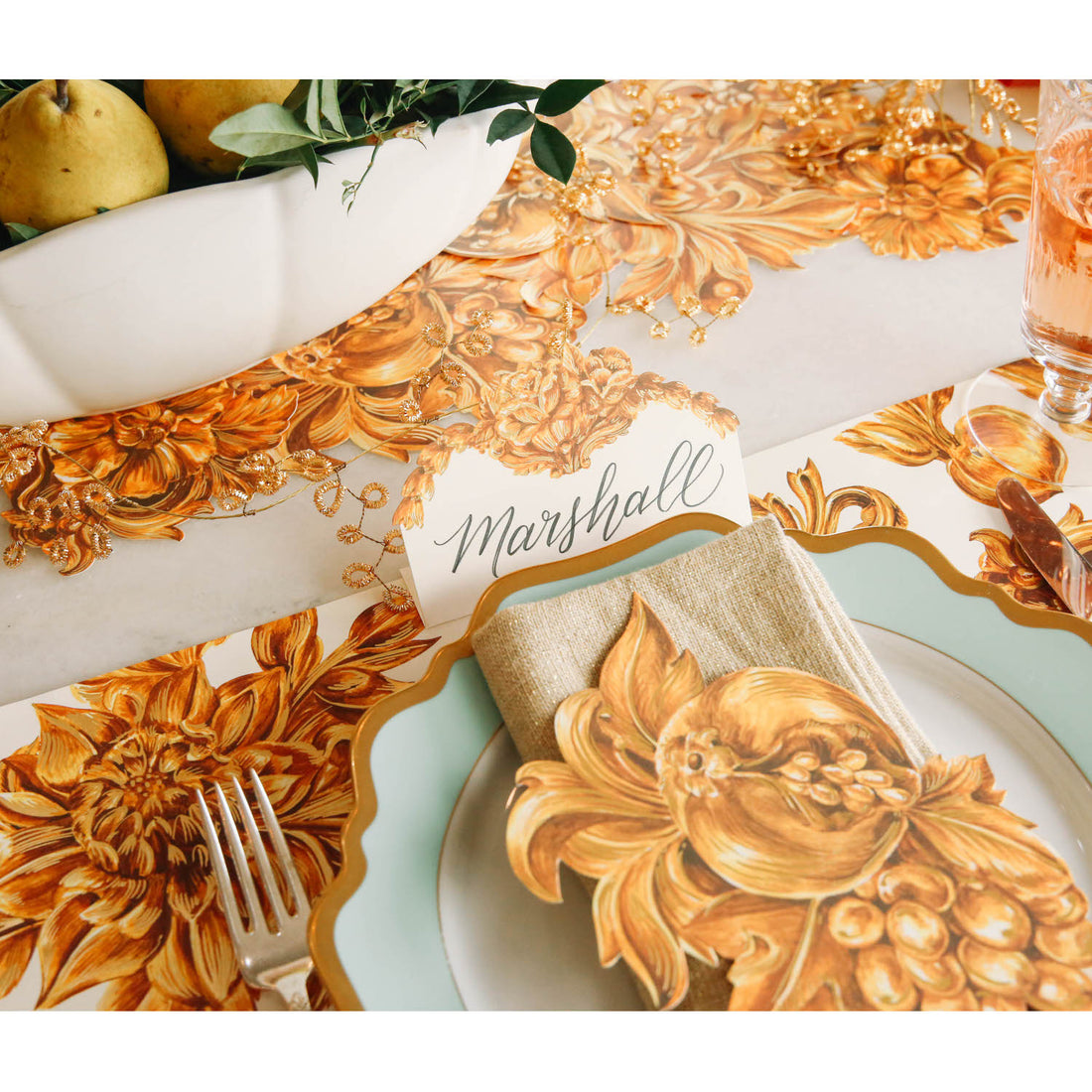 An elegant gold and white place setting featuring a Gold Flora Place Card labeled "Marshall" standing behind the plate.