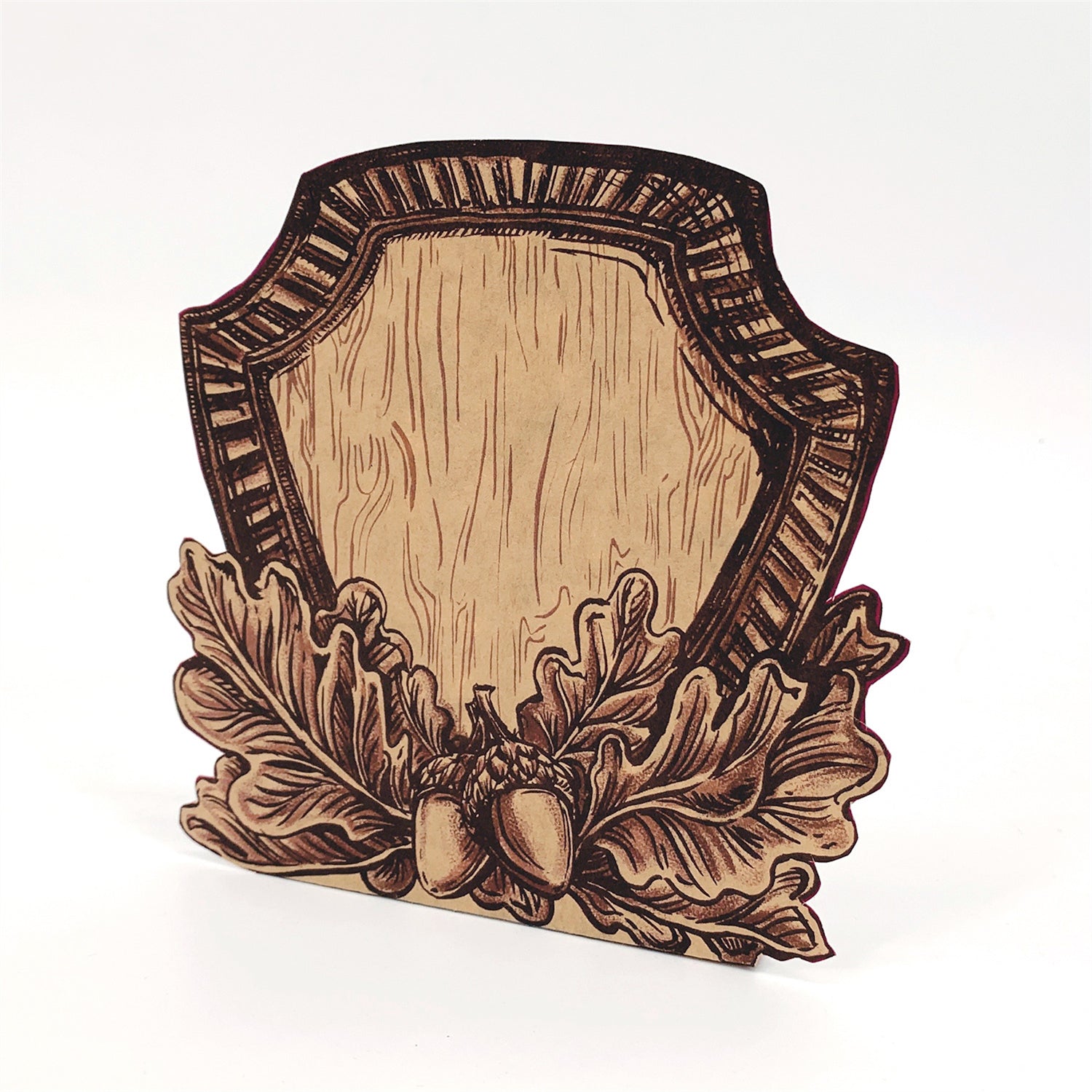 A die-cut, freestanding kraft paper place card featuring deep brown linework of a wooden shield adorned with oak leaves and acorn across the bottom.
