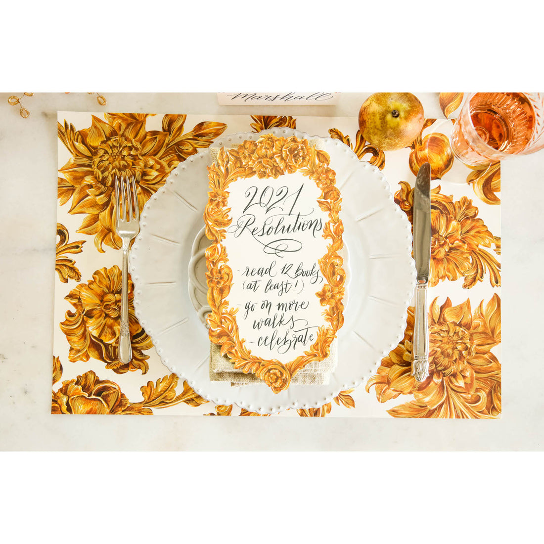 Top-down view of an elegant gold and white place setting featuring a Gold Flora Table Accent with some New Year Resolutions written on it in a beautiful script.