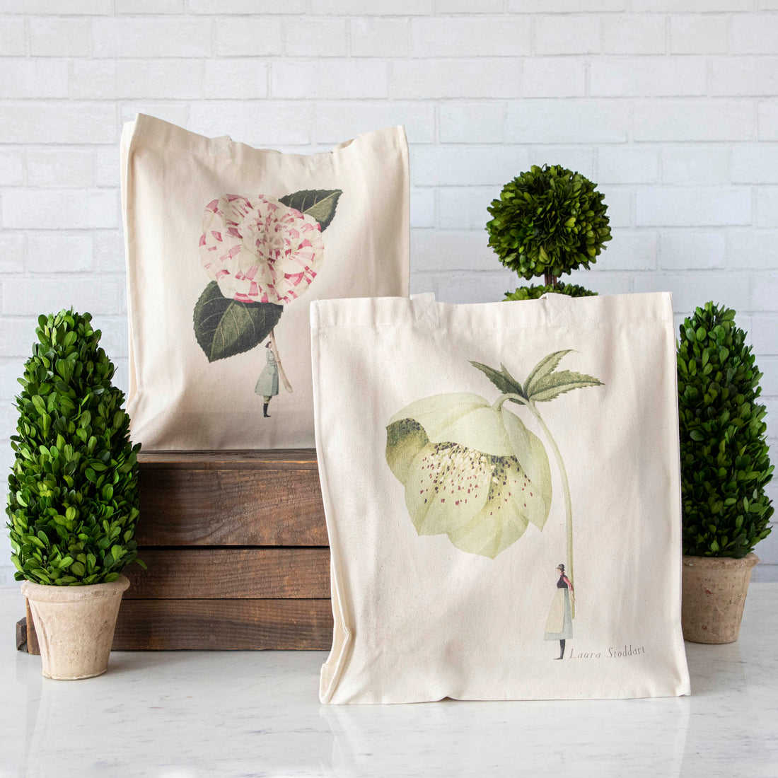 Two Hester &amp; Cook heavyweight canvas tote bags with floral prints leaning against wooden crates beside potted topiary plants.