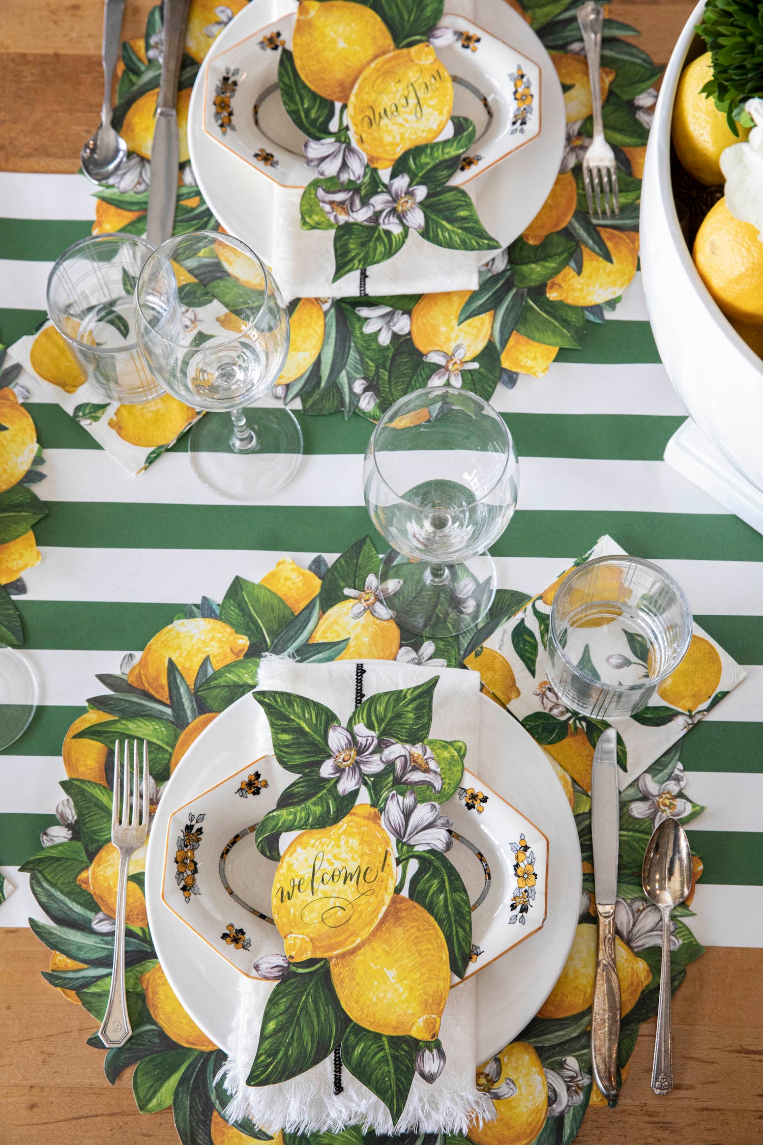 A table setting with Die-cut Lemon Wreath Placemats by Hester &amp; Cook on a green and white striped tablecloth.