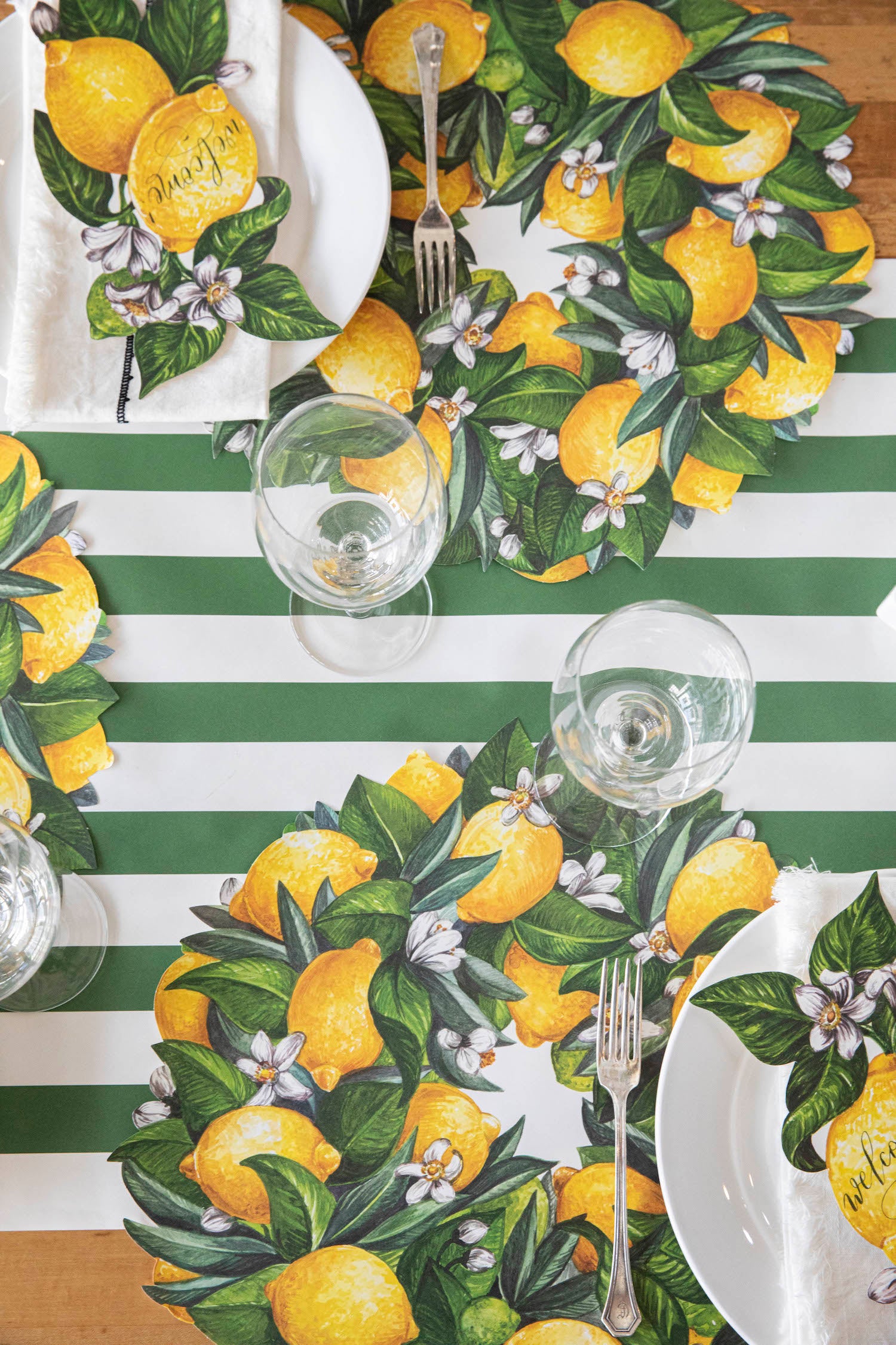A table setting with Die-cut Lemon Wreath Placemat by Hester &amp; Cook, lemons, and green leaves.