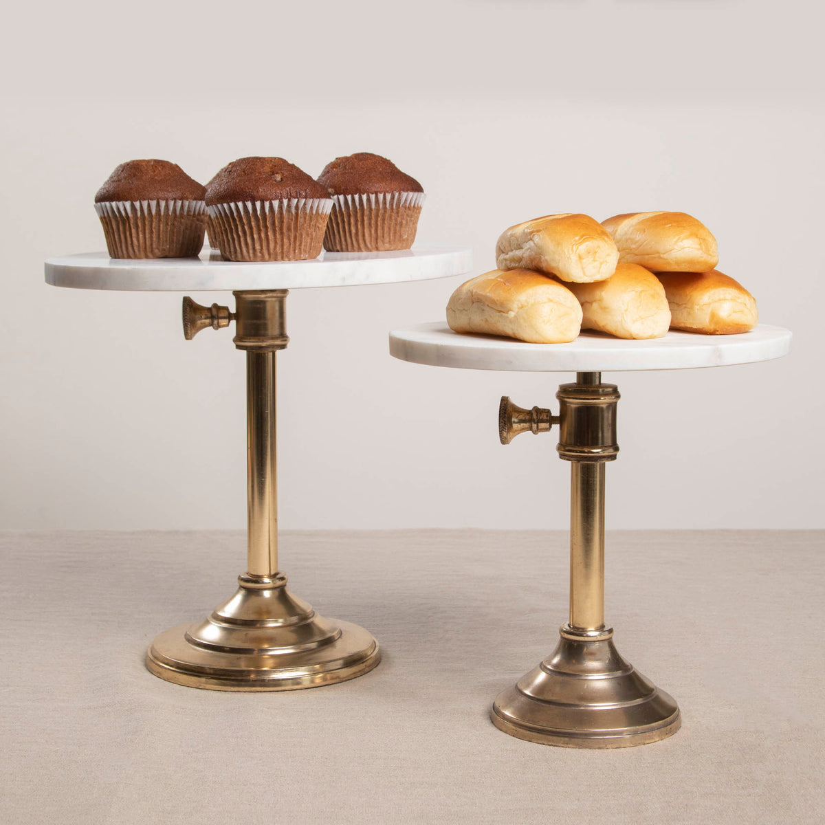 Two adjustable cake plates with marble tops and antique brass stands, one expanded to a larger size displaying muffins, and the other closed showing rolls.
