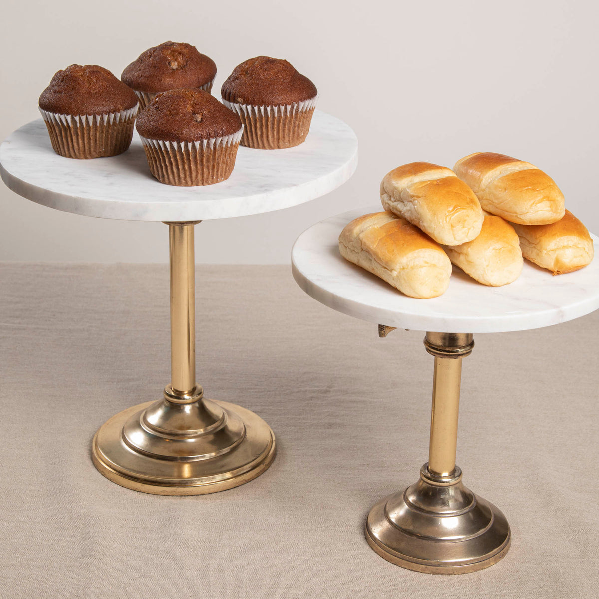 Two adjustable cake plates with marble tops and antique brass stands, one expanded to a larger size displaying muffins, and the other closed showing rolls.