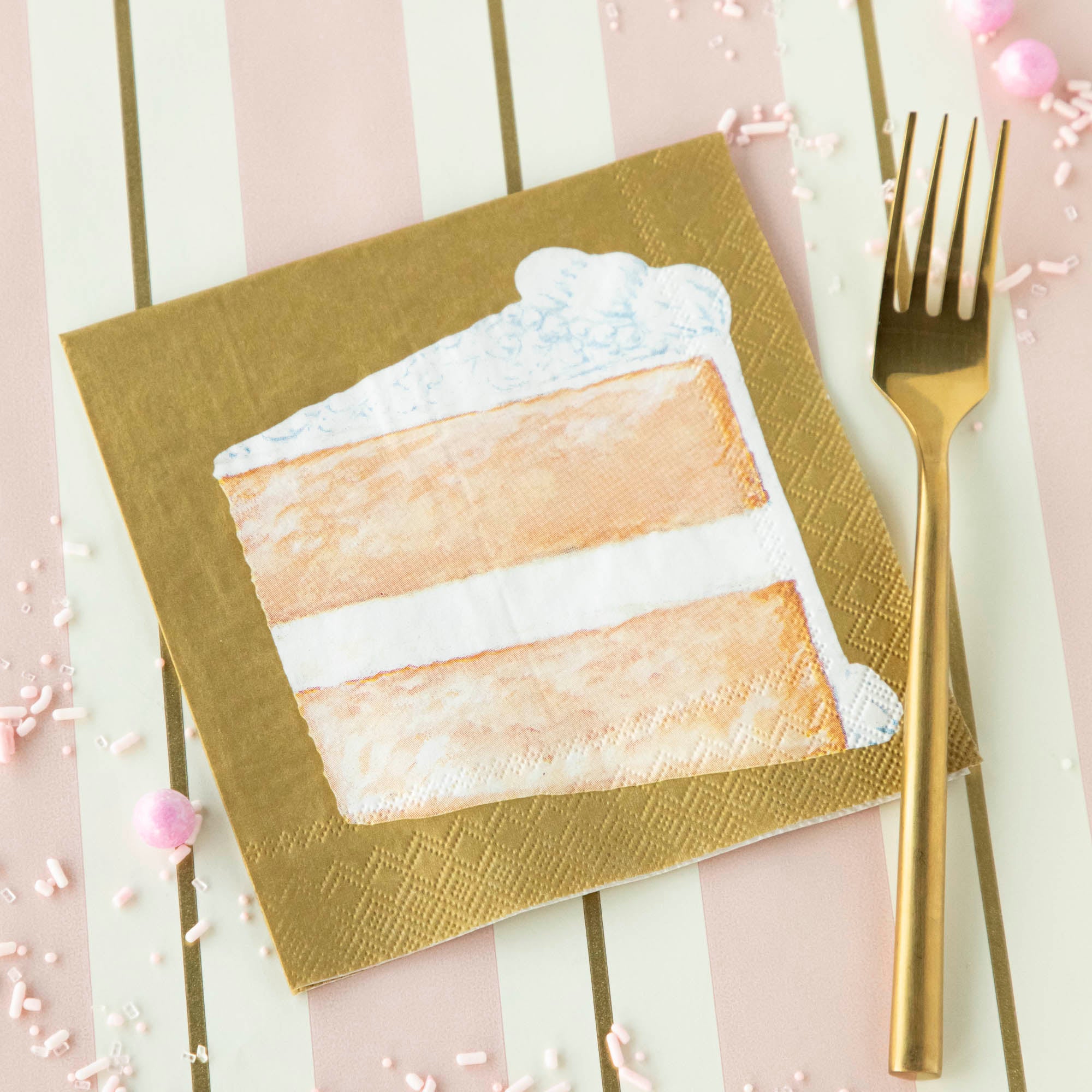 A Sliced Cake Cocktail Napkin on a pink table runner with a gold fork.